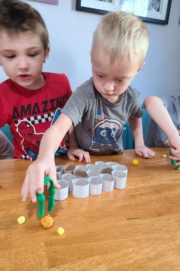 Two young boys play at a table, using green tongs to pick up small yellow pom-poms near a row of white cardboard cylinders—a hands-on activity perfect for lesson plans on bees.