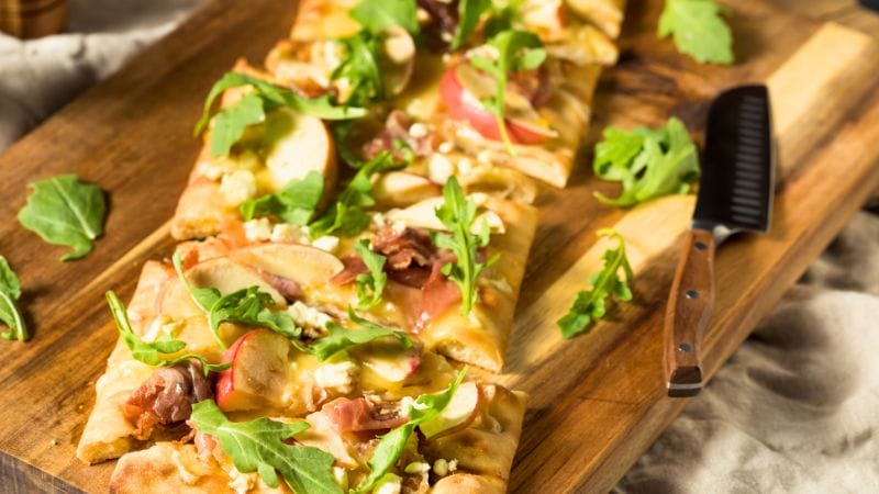 Rectangular flatbread pizza topped with arugula, apple slices, prosciutto, and crumbled cheese—a creative dinner idea with lunch meat—served on a wooden board next to a knife.