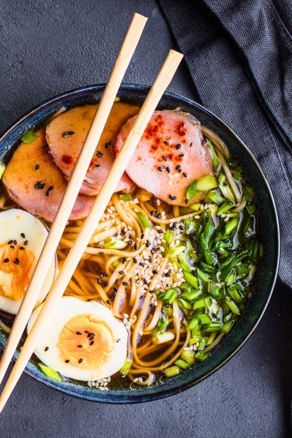 A bowl of ramen featuring sliced lunch meat, soft-boiled egg halves, noodles, chopped greens, sesame seeds, and chopsticks resting on top—perfect for creative dinner ideas with lunch meat.