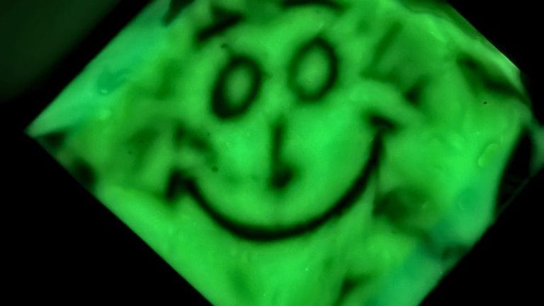 A blurry, green-tinted image of a smiling face drawn with simple lines, as if seen through a Glow in the Dark Sensory Bag. The face appears distorted, with visible smudges and water droplets.