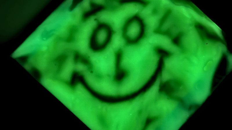 A blurry, green-tinted image of a smiling face drawn with simple lines, as if seen through a Glow in the Dark Sensory Bag. The face appears distorted, with visible smudges and water droplets.