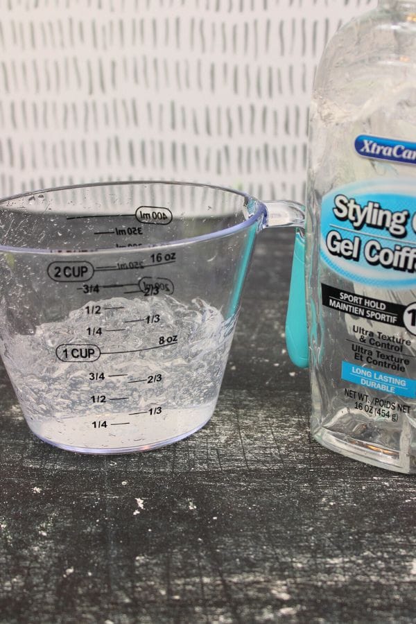 A clear measuring cup with gel residue next to a partially visible bottle of XtraCare Styling Gel sits on a dark, textured surface—perfect for prepping a Glow in the Dark Sensory Bag—against a patterned background.