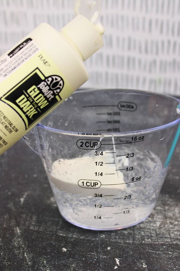 A bottle of glow-in-the-dark glue is being poured into a measuring cup of clear liquid on a black surface, the first step in creating a Glow in the Dark Sensory Bag.