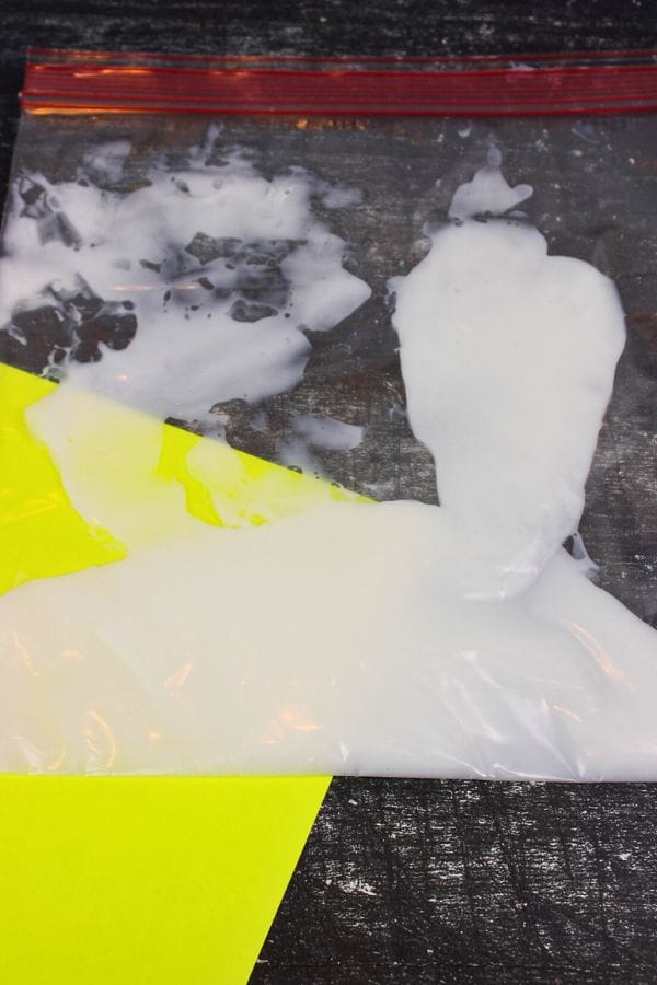 A Glow in the Dark Sensory Bag filled with white liquid rests on a dark surface, with a yellow paper partially tucked underneath.