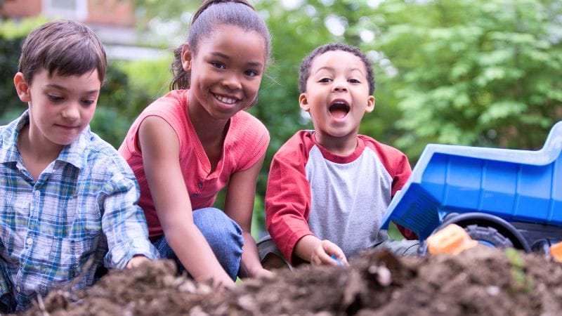 Three children play outdoors in the Home Daycare backyard, with one smiling at the camera, another laughing, and a toy dump truck to the right.
