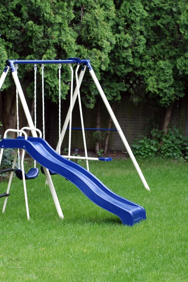 A blue and white playground set with a slide and swings stands on green grass in a home daycare backyard, with trees and a fence in the background.