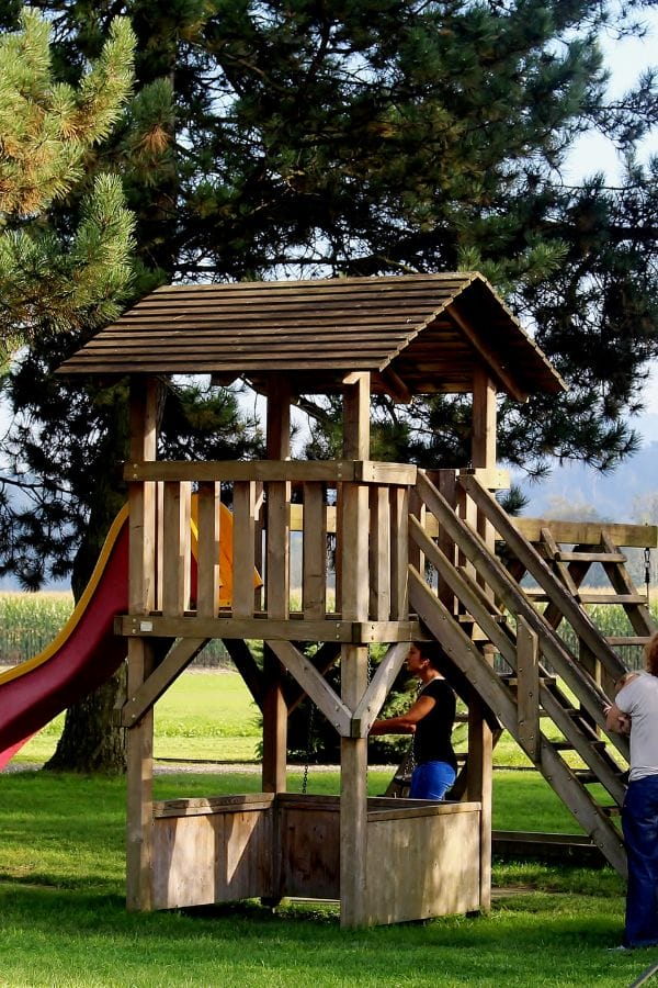 A wooden playground structure with stairs, a roofed platform, and a red slide stands on grass in a Home Daycare Backyard; two people are nearby and large trees fill the background.