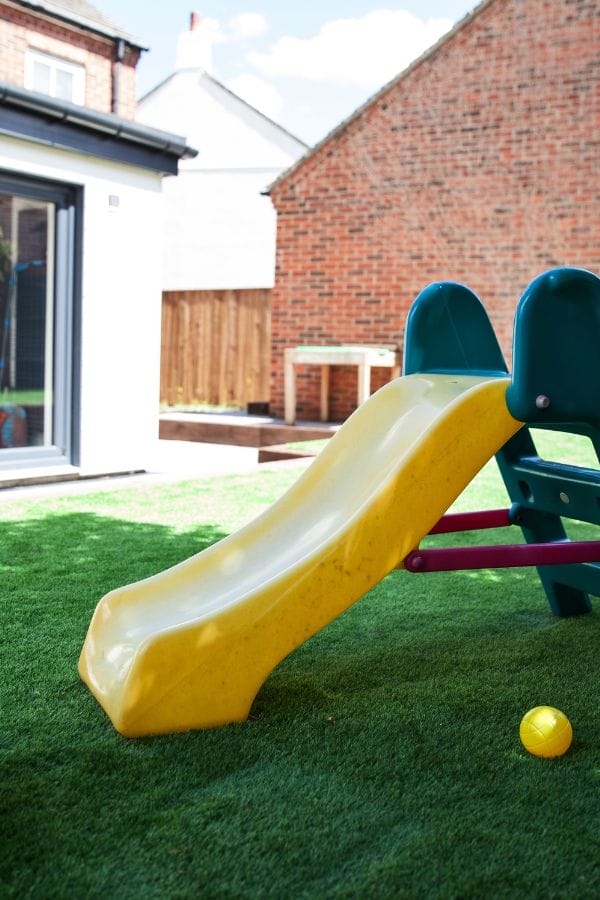 A yellow plastic slide on artificial grass in a home daycare backyard, with a yellow ball on the ground nearby and brick buildings in the background.