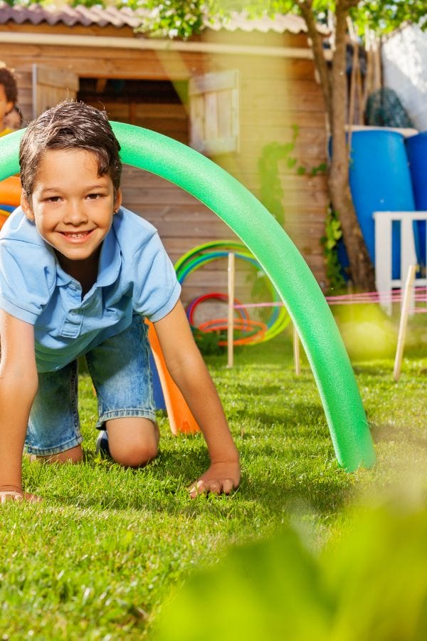 A young boy in a blue shirt and denim shorts crawls under a green foam pool noodle on the grass in a Home Daycare Backyard, smiling, with playground equipment visible in the background.