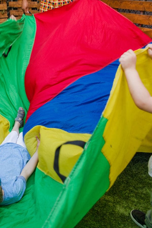 Children in a home daycare backyard hold and play with a large, colorful parachute on the grass, while one child lies underneath it.