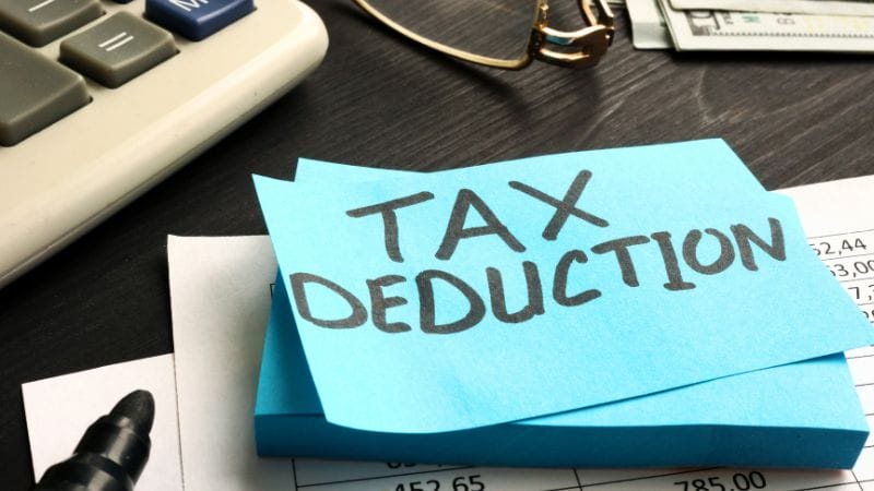 A stack of blue sticky notes with "TAX DEDUCTION" written on top, placed on a desk with a calculator, paperwork, eyeglasses, and cash nearby—perfect for organizing home daycare taxes.