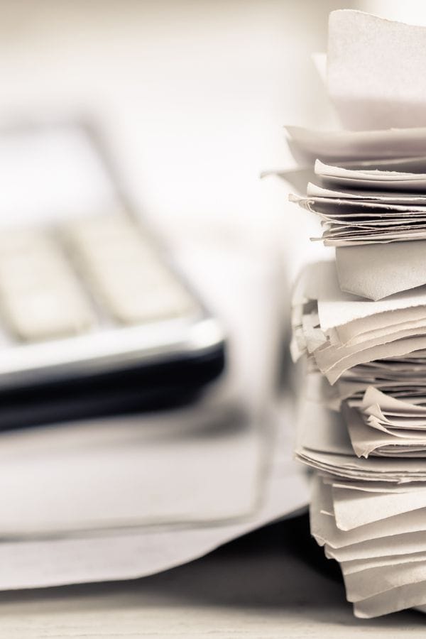 A close-up of a tall stack of papers, possibly related to home daycare taxes, sits next to a blurred calculator on a desk.