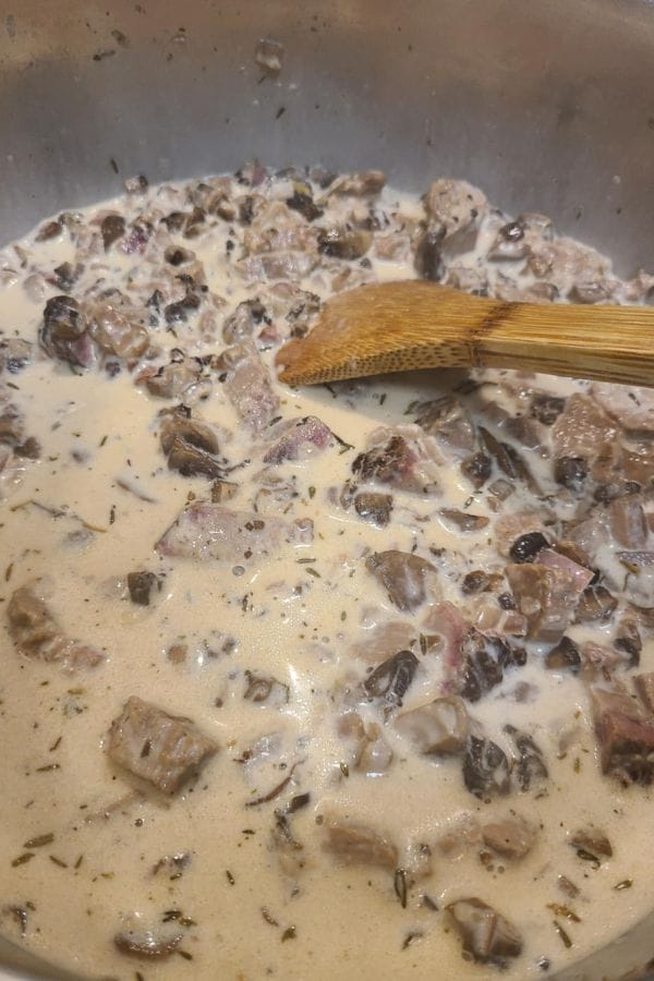 Chunks of beef and mushrooms simmer in a creamy sauce inside a large pan, making this Leftover Brisket Pasta with Mushrooms a hearty, comforting meal—complete with a wooden spatula resting on the delicious mixture.