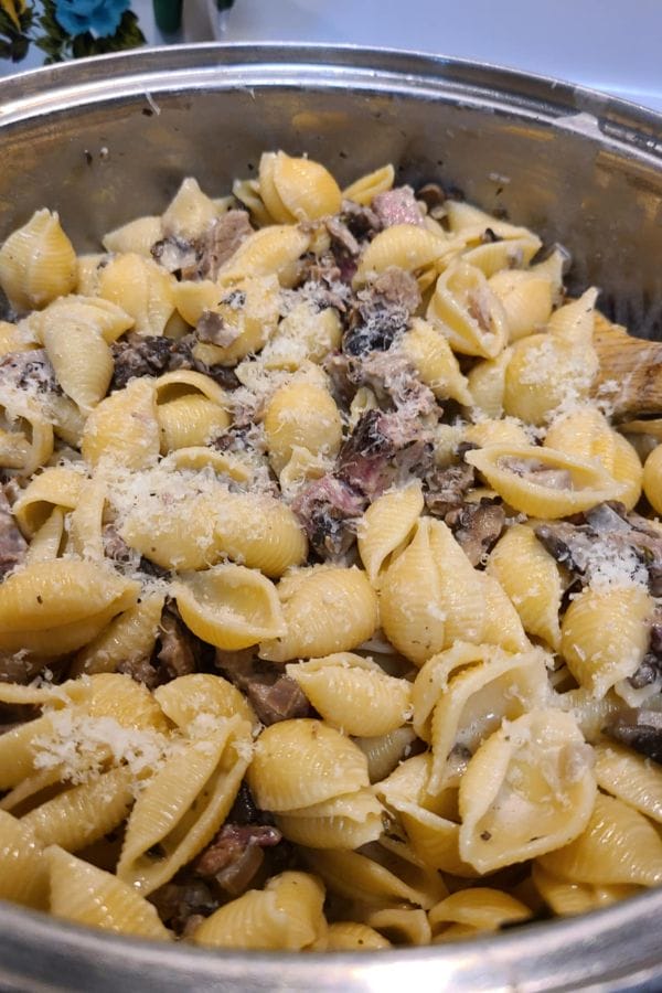 A stainless steel pot filled with shell pasta, sliced mushrooms, leftover brisket pieces, and grated cheese on top—a comforting Leftover Brisket Pasta with Mushrooms.