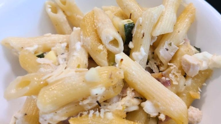 A close-up of Penne Tuna Pasta Bake, featuring cooked penne pasta mixed with cheese, herbs, and small pieces of vegetables in a white bowl.