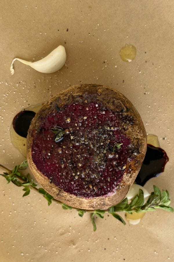 A seasoned roasted beet next to a clove of garlic, fresh herbs, and a drizzle of dark sauce on brown parchment paper highlights the rich flavors of roasted beets in this vibrant presentation.