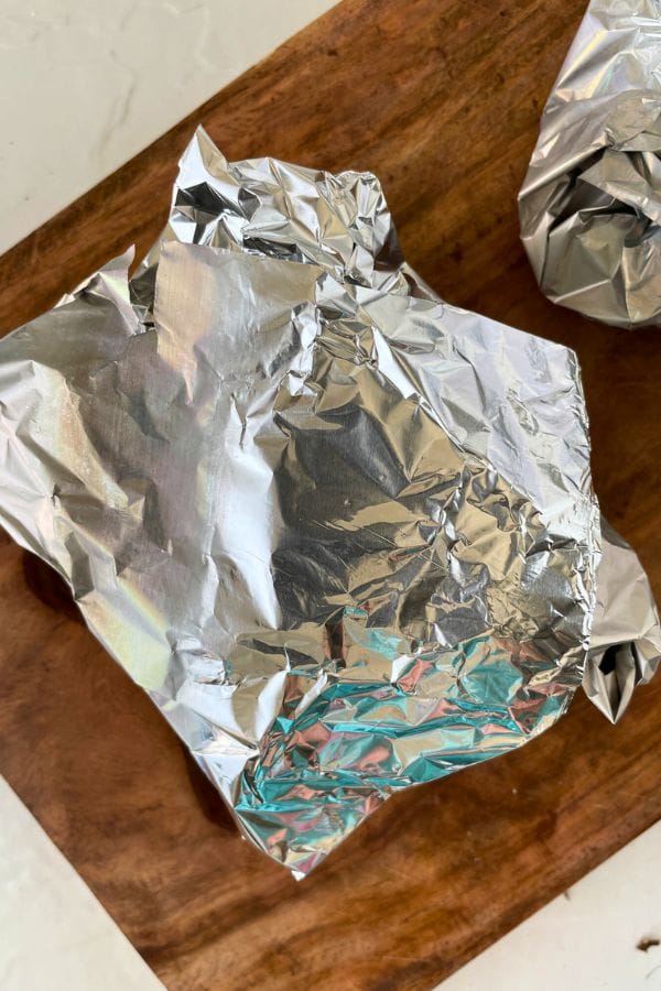 A piece of food, possibly roasted beets, wrapped in crumpled aluminum foil sits on a wooden surface, with part of another foil-wrapped item visible in the corner.