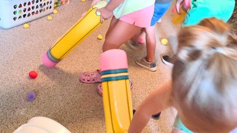 Children enjoy a lively Pencil Party indoors, using large inflatable pencils to push colorful plastic balls across the carpeted floor. A laundry basket and scattered balls add to the festive atmosphere in the background.