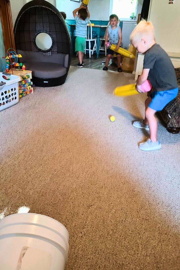 Three young children play baseball indoors with plastic bats and a ball on a carpeted floor while hosting a lively Pencil Party; toys and furniture are visible in the background.