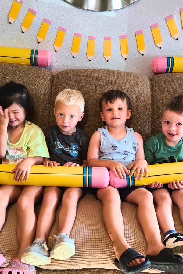 Four young children sit on a couch holding large inflatable pencils at a Pencil Party, with paper pencil decorations displayed on the wall behind them.