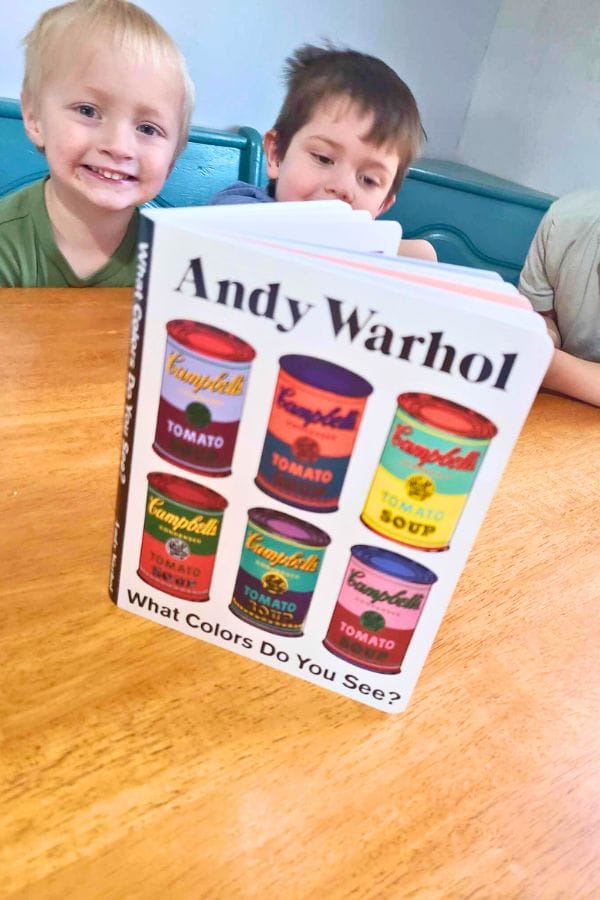 Two children sit at a table behind a book titled "Andy Warhol: What Colors Do You See?"—a perfect companion for lesson plans on Andy Warhol, with its colorful Campbell's soup cans featured on the cover.