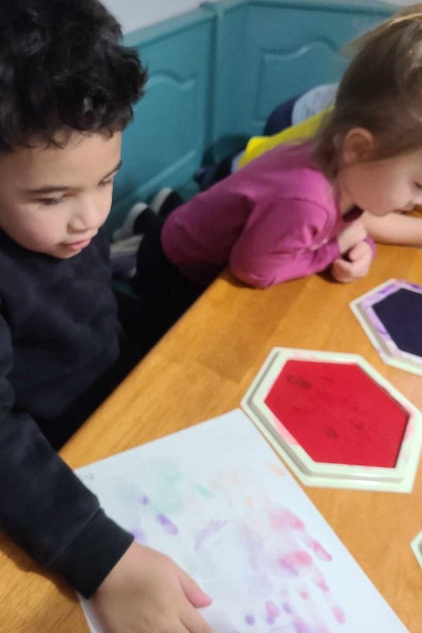 Two young children sit at a table, looking at a piece of paper with colorful smudges and hexagonal shapes nearby—perfect inspiration for lesson plans on Andy Warhol.