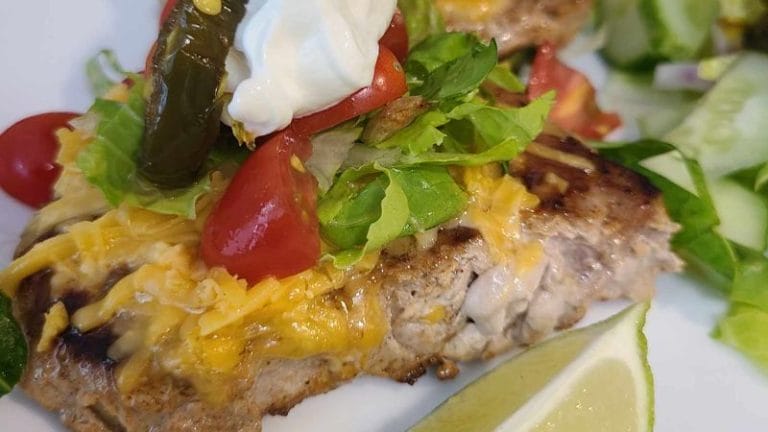 Close-up of Taco Pork Chops topped with shredded cheese, lettuce, tomatoes, jalapeño, sour cream, and served with a lime wedge and sliced cucumbers.