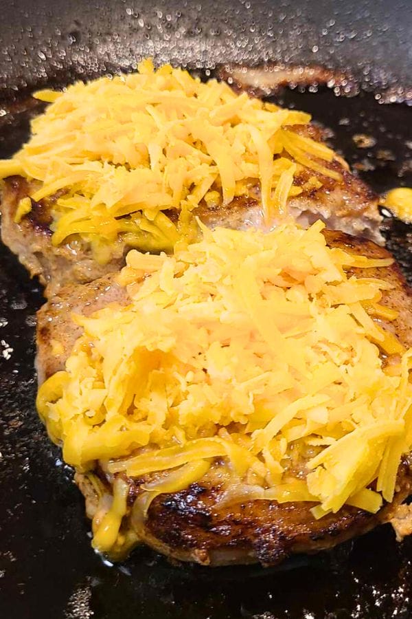 Two beef patties cooking in a skillet, each topped with a generous layer of shredded cheddar cheese, perfect for pairing with flavorful Taco Pork Chops.