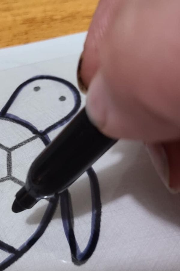 A hand draws the outline of a turtle with a black marker on white paper, creating a perfect design for a School Shrinky Dink project.