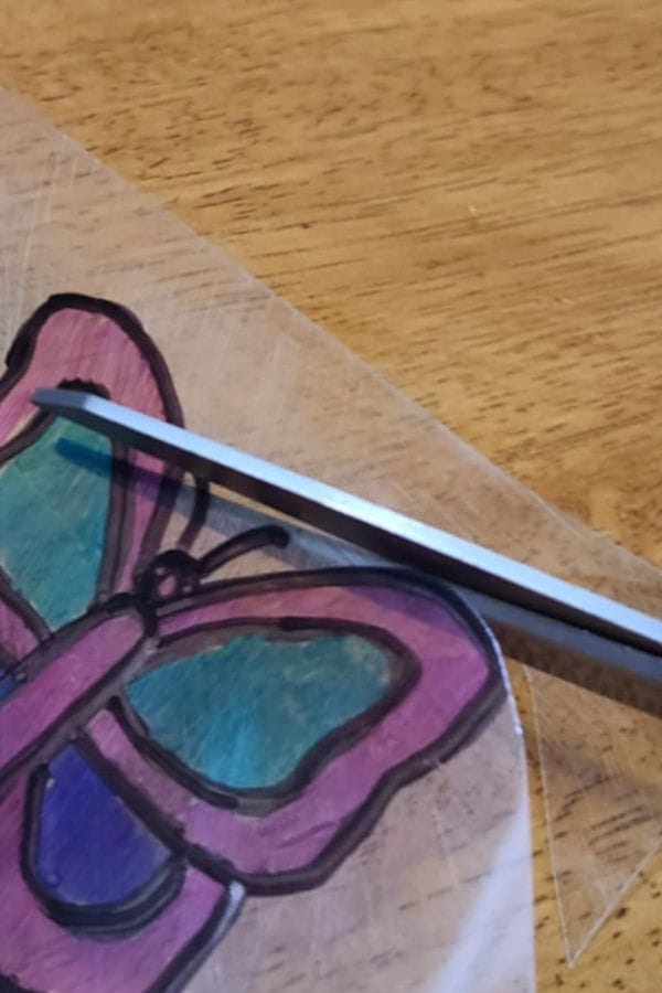 A pair of scissors is cutting out a hand-drawn and colored butterfly on a School Shrinky Dink sheet, placed on a wooden surface.