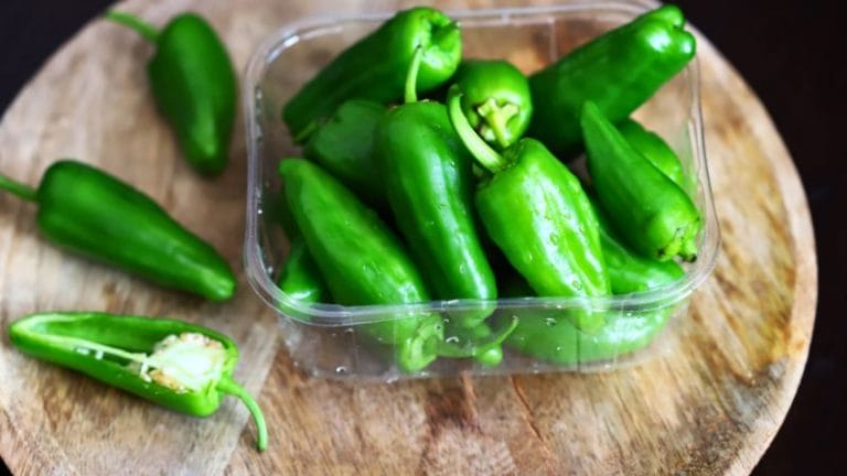 A clear plastic container filled with fresh green jalapeño peppers sits on a round wooden cutting board, tempting anyone interested in how to grow jalapenos at home. A few peppers are placed beside it for added freshness.