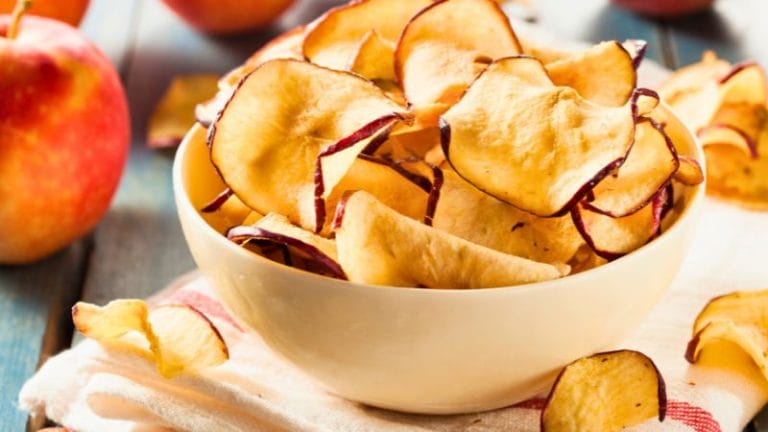 A white bowl filled with dried apple chips sits on a cloth, with whole apples and more chips scattered around on a wooden surface—an inviting way to store dehydrated food for healthy snacking.