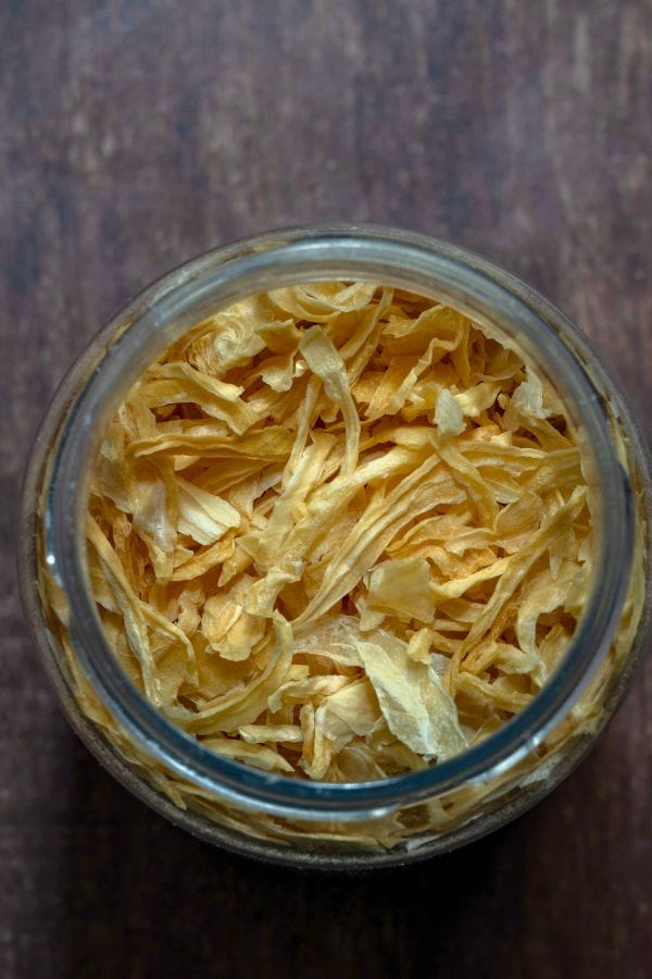 A glass jar filled with dried onion flakes, viewed from above, is placed on a dark wooden surface—an ideal way to store dehydrated food and keep your pantry organized.