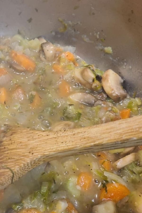 A close-up of a pot with chopped mushrooms, carrots, celery, and herbs being stirred with a wooden spoon—perfect for starting a hearty Leftover Brisket Pot Pie.