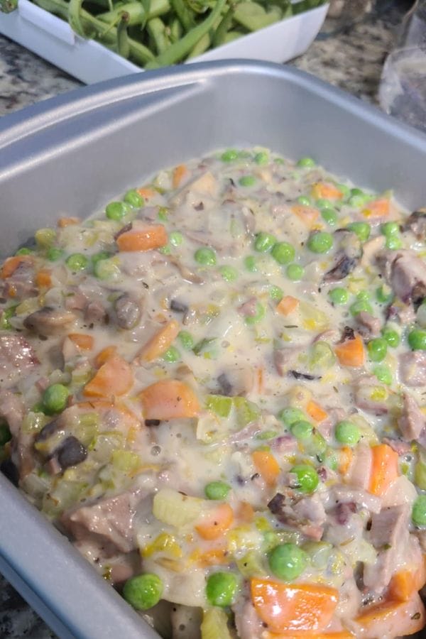 A baking dish filled with a mixture of diced vegetables, peas, and leftover brisket in a creamy sauce, ready to be cooked as a delicious Leftover Brisket Pot Pie. A container of green beans is visible in the background.
