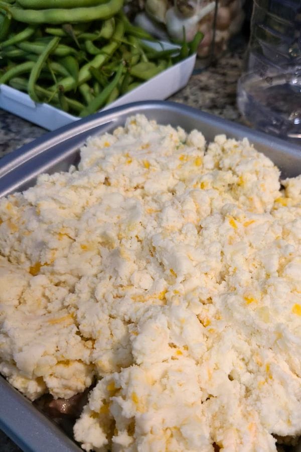 A baking dish filled with an unbaked layer of biscuit dough, ready to become a savory Leftover Brisket Pot Pie, with fresh green beans and mushrooms in the background.