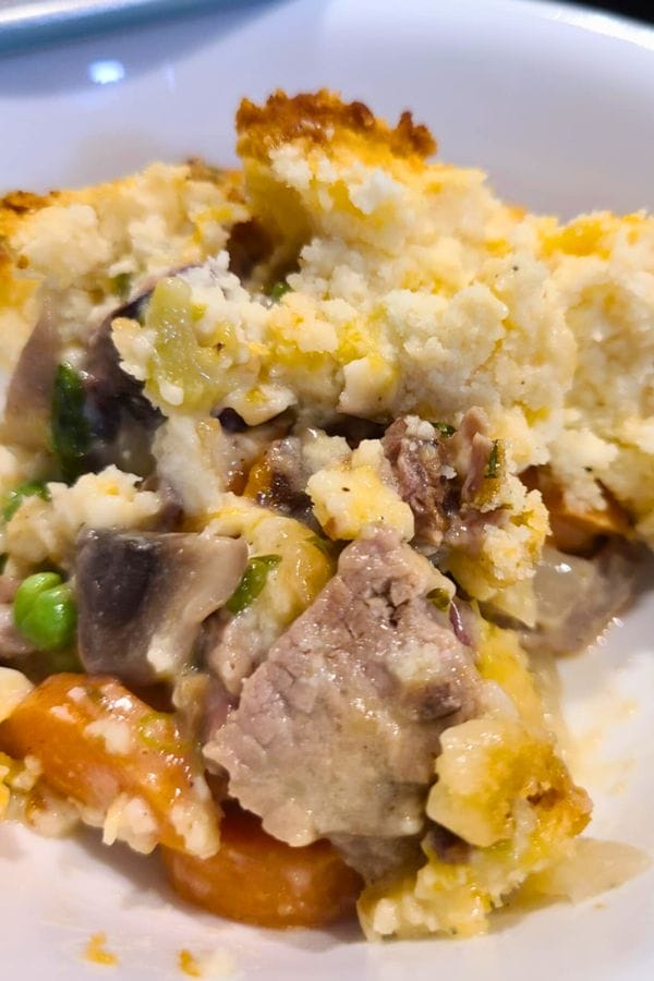 A close-up of a serving of Leftover Brisket Pot Pie and vegetable casserole, topped with a layer of baked, golden biscuit crust in a white bowl.
