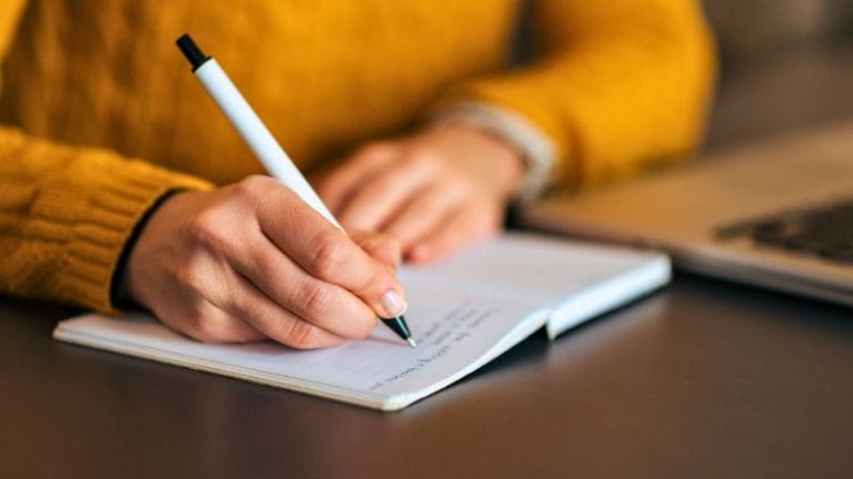 A person wearing a yellow sweater writes in a notebook with a pen, next to a laptop on a desk, possibly researching Oklahoma daycare licensing regulations.