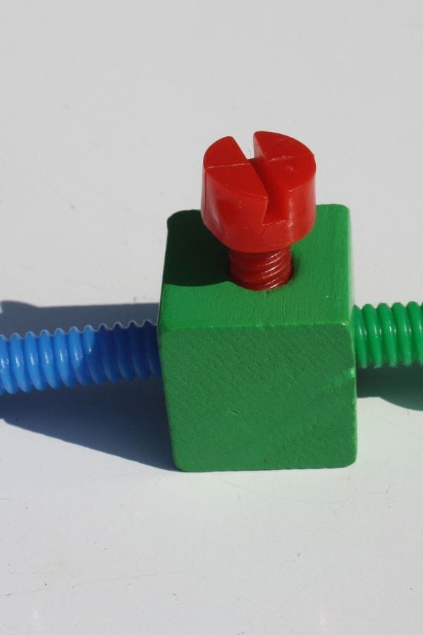 A red plastic screw is inserted into a green wooden block with blue and green flexible tubes extending from opposite sides against a white background, illustrating materials that could comply with Oklahoma daycare licensing regulations.