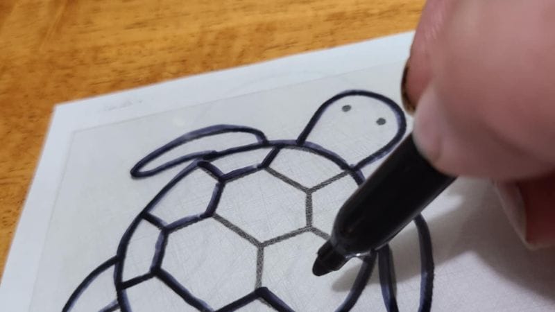 A hand uses a black marker to trace the outline of a turtle drawing, inspired by Fall Shrinky Dink Patterns, on a sheet of paper placed on a wooden surface.
