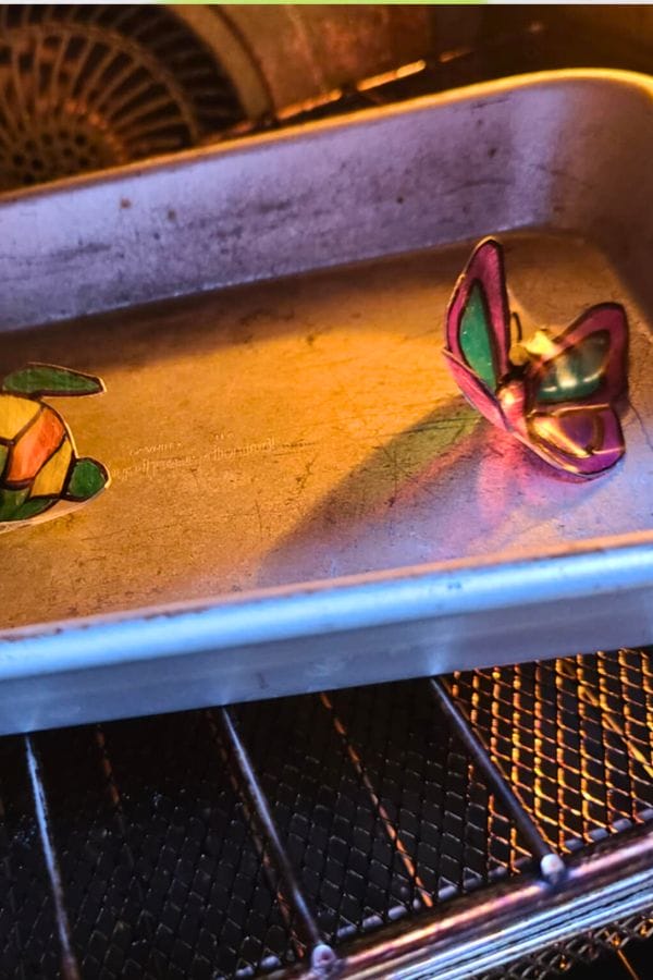 A metal baking tray with Fall Shrinky Dink Patterns—colorful plastic pieces—is placed in an oven, with the designs starting to curl from the heat.