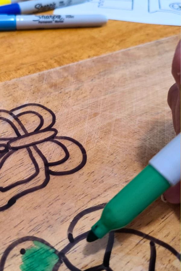 A hand uses a green marker to color a drawing on a wooden surface, with Sharpie markers, Fall Shrinky Dink Patterns, and paper visible in the background.