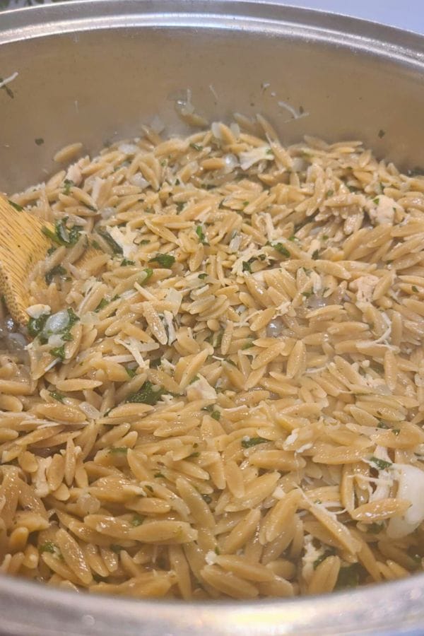 A pot filled with cooked tuna and orzo pasta mixed with herbs and shredded cheese, being stirred with a wooden spoon.