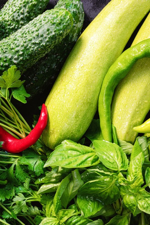 Assorted fresh vegetables and herbs, including cucumbers, yellow zucchinis, red and green chili peppers, parsley, and basil—popular companion plants for peppers that thrive together in any garden.