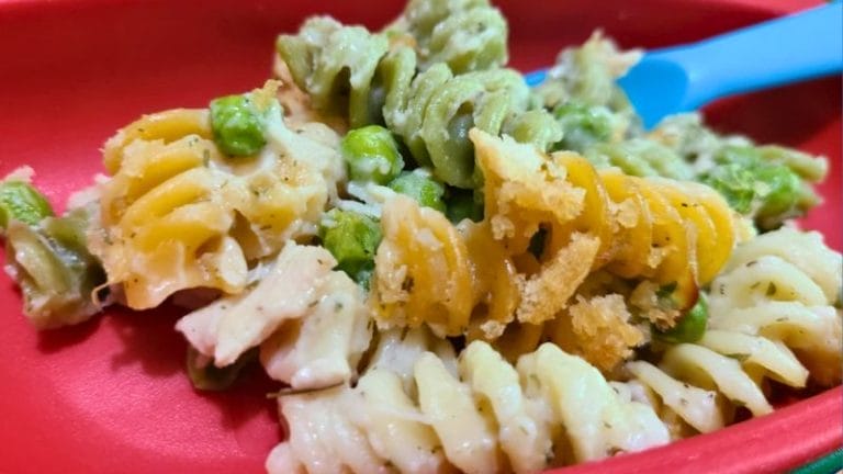 Close-up of tri-color rotini pasta with peas and a creamy, dairy free tuna casserole sauce, served in a red dish with a blue utensil.