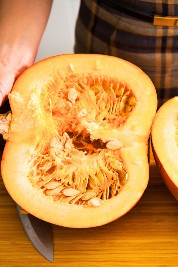 A hand holding a halved pumpkin showing its seeds and fibrous interior, with a knife on a wooden surface below.