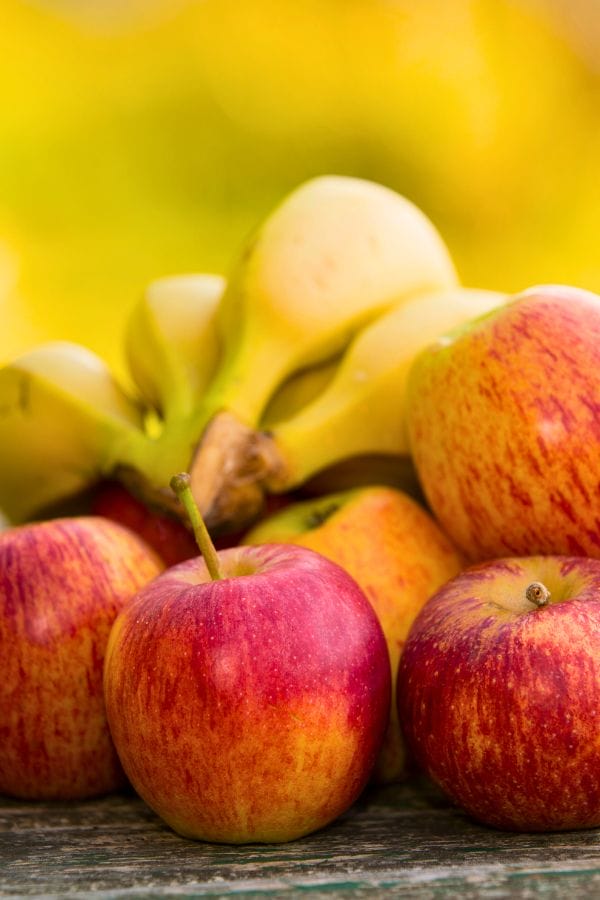 A bunch of bananas and several red-yellow apples placed together on a wooden surface with a blurred yellow background.