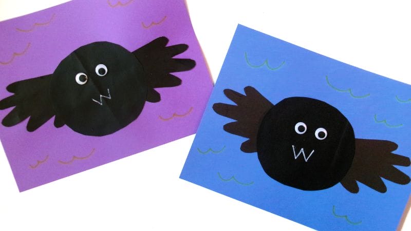 Two handmade Handprint Bat paper crafts with black circular bodies and handprint wings are displayed on purple and blue backgrounds, adorned with simple squiggly line decorations.