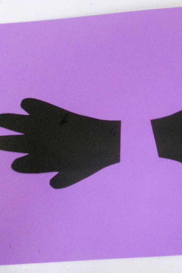 Two black paper mittens resembling a handprint bat are placed on a purple background, facing away from each other.