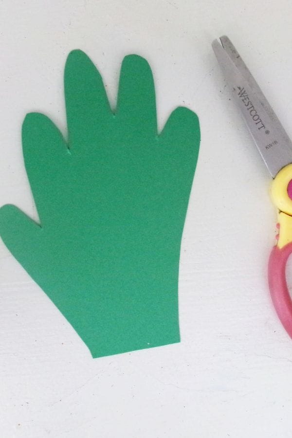Green paper cut in the shape of a hand sits next to a pair of scissors with red and yellow handles on a white surface, perfect for creating a Handprint Pumpkin Craft.